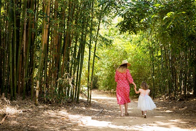 Lady in a red dress and a wide brimmed sun hat walking along a bamboo-lined path holding hands with a young girl in a white dress.