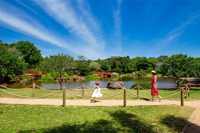 Lady wearing a red long dress and a wide brimmed sundress walking along a path with a young girl in a white dress. The young girl is doing a swirl. Lake in the background with red traditional style bridges crossing over.