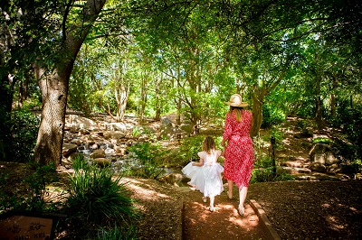 Lady in a red dress and wide brimmed sun hat walking with a young girl wearing a white dress. A shaded leafy section of the park with water running over rocks.