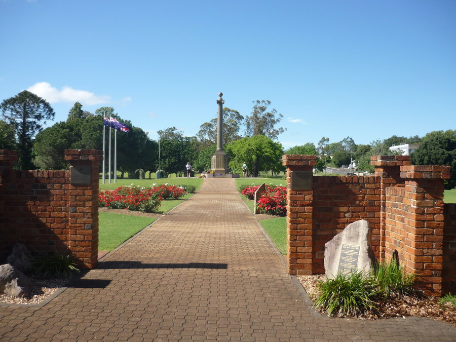 Photo of Mothers' Memorial. Paved path leading to monument in the middle of a grassed area. Three flagpoles to the left of the monument.