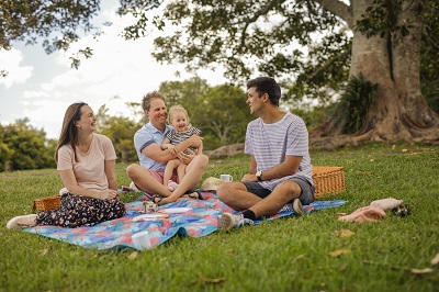 Two men, a lady and a young girl enjoying a picnic on the open grass at the Cedar Block day-use area. Group sitting on a picnic rug smiling and laughing.