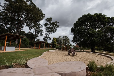 Children's nature play area at the Highfields Peacehaven Botanic Park. Large sand play area with sculpture in the middle, surrounded by picnic shelters. 